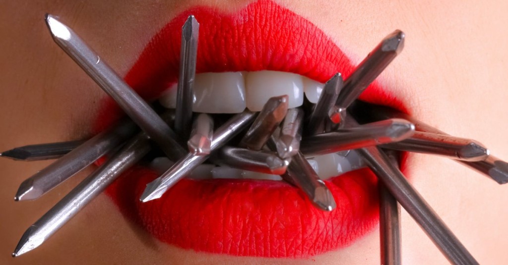 A close up of a pair of red lips and parted teeth holding metal pieces