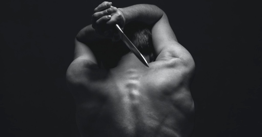 A muscular, shirtless man with his back towards the camera and a knife raised with both hands at his neck level