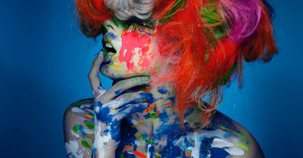 The bust shot of suggestive pose of a young woman covered in different colored paint with a a multicolored wig on her head