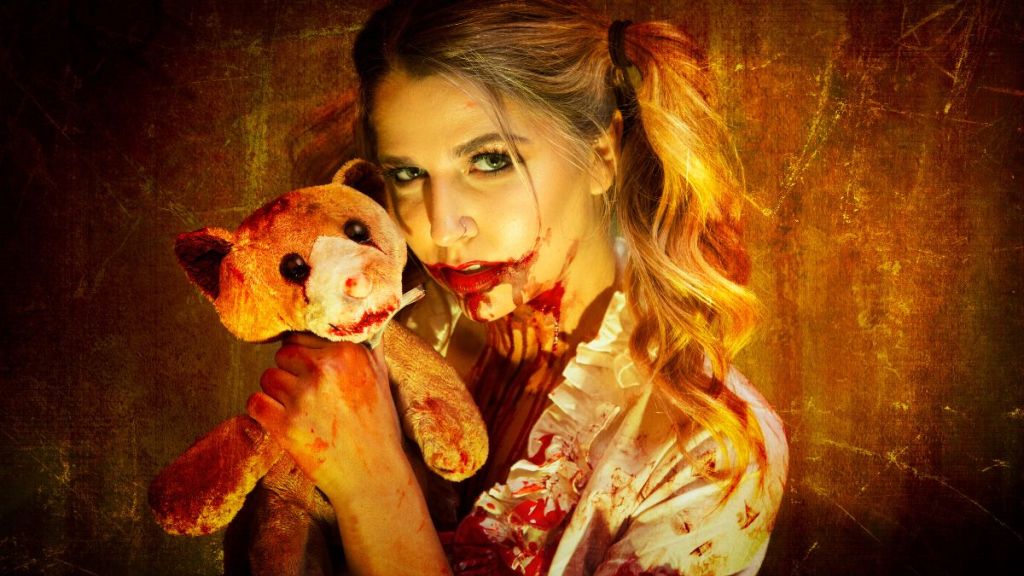 A young woman with blood smeared on her face and clothes holding a battered and bloody teddy bear