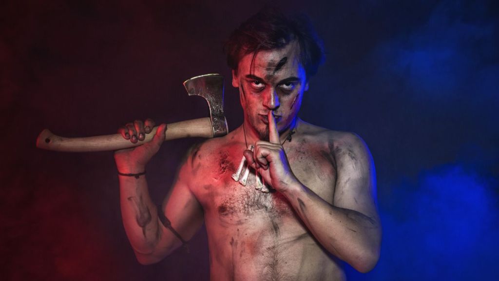 A bare chested young man with black marking on his body and holding an axe up in one hand and the index finger of his other hand is placed on his lips