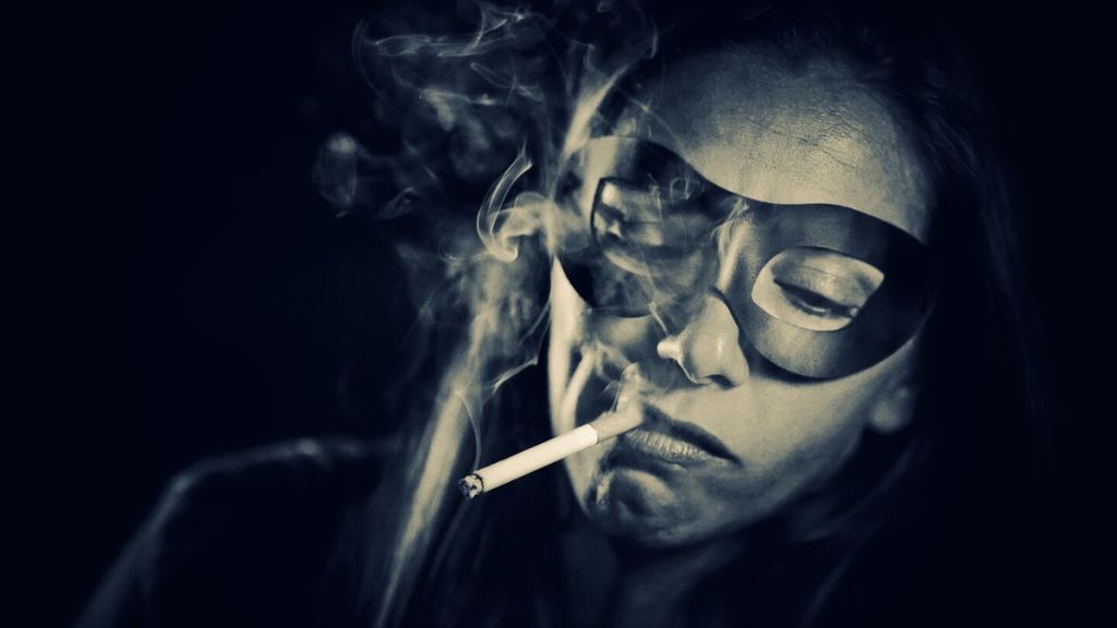 The close up black and white image of a woman wearing a superhero eye mask and smoking a cigarette