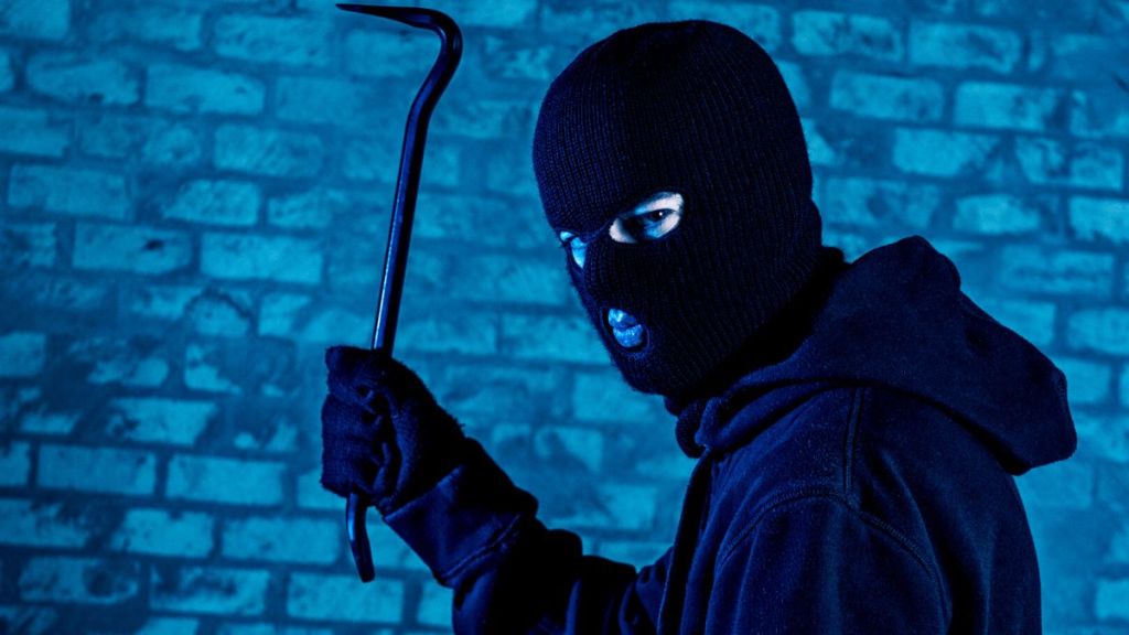 A woman in a ski mask looking sideways and holding a crowbar in a dark alley