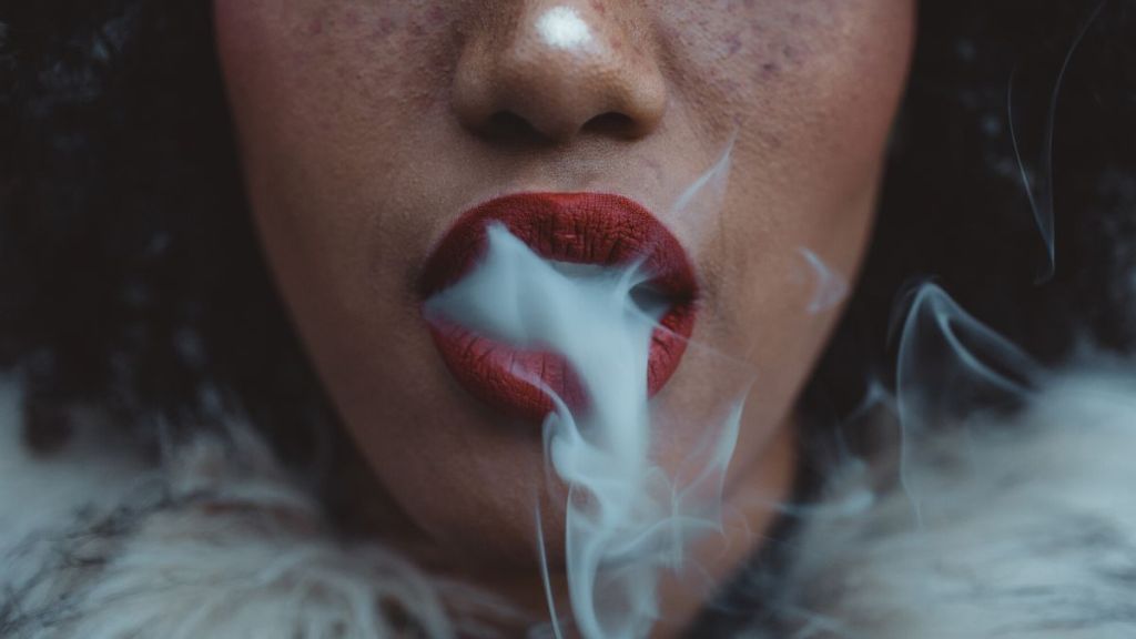The close up of a dark-skinned woman's red lipstick mouth blowing a thick plume of smoke