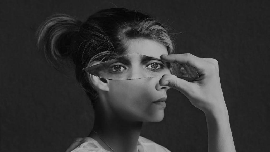 The black and white side profile of a woman's face holding up a broken shard of glass against her face with the reflection of two eyes in it