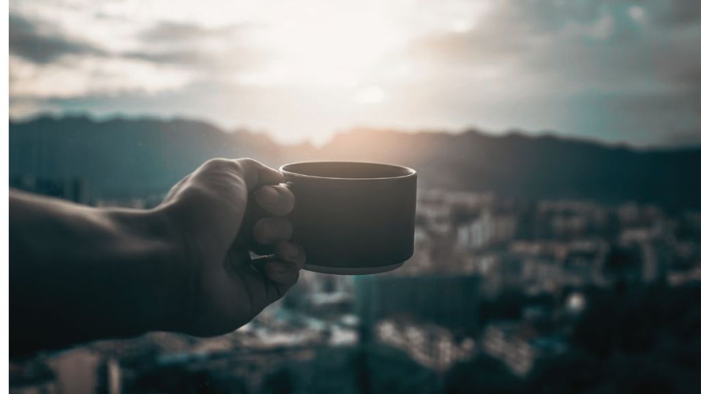 A man's hand holding out a cup of coffee in the foreground with a view of a city in the distant background