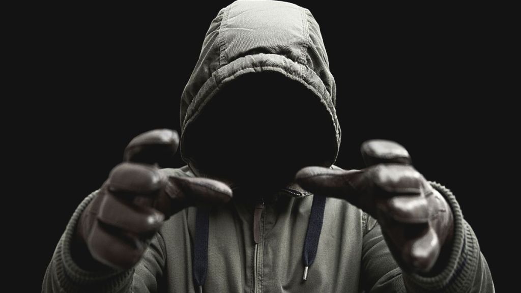A dark figure in a black hoodie with gloved hands reaching out as if to choke someone