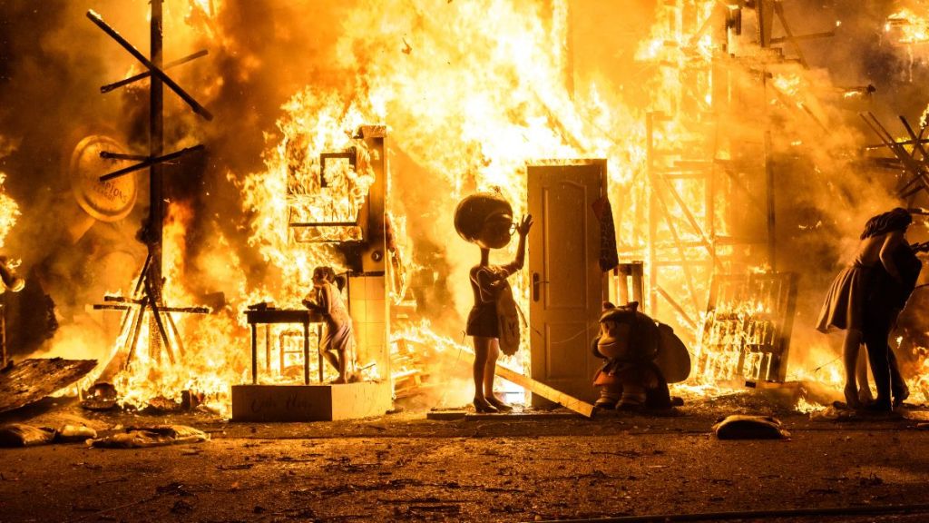 An animated scene in which there is a raging fire that's burning a house and a young girl stands at the door and is about to leave