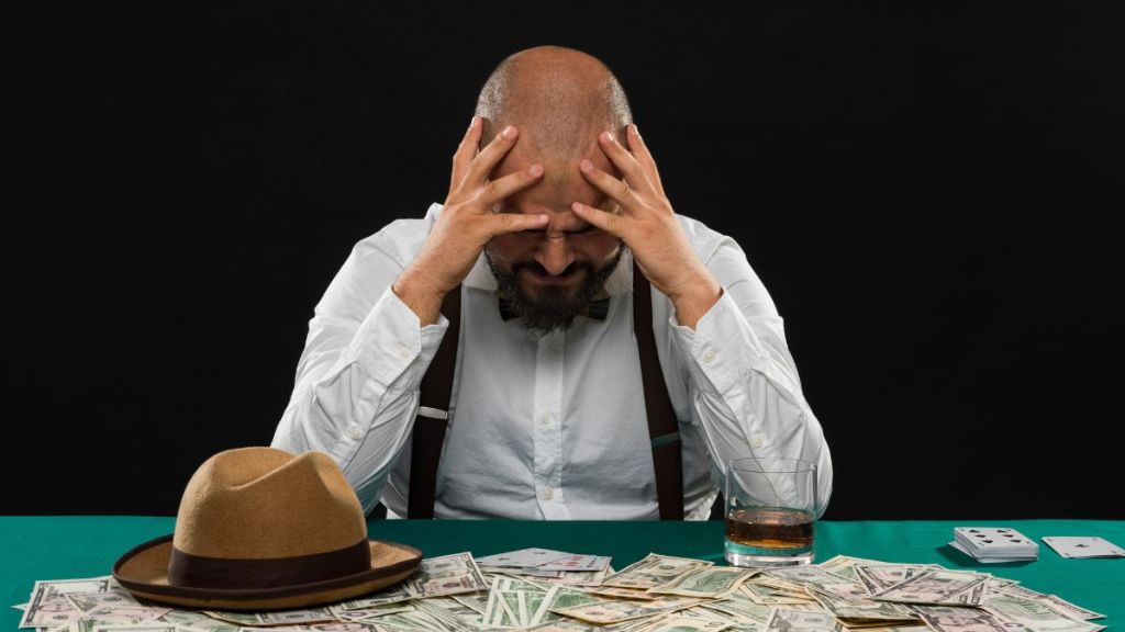 A bald, bearded man in a white shirt and suspenders holding his head and looking down in pain as his hat sits next to him and many dollar notes are spread in front of him on the table