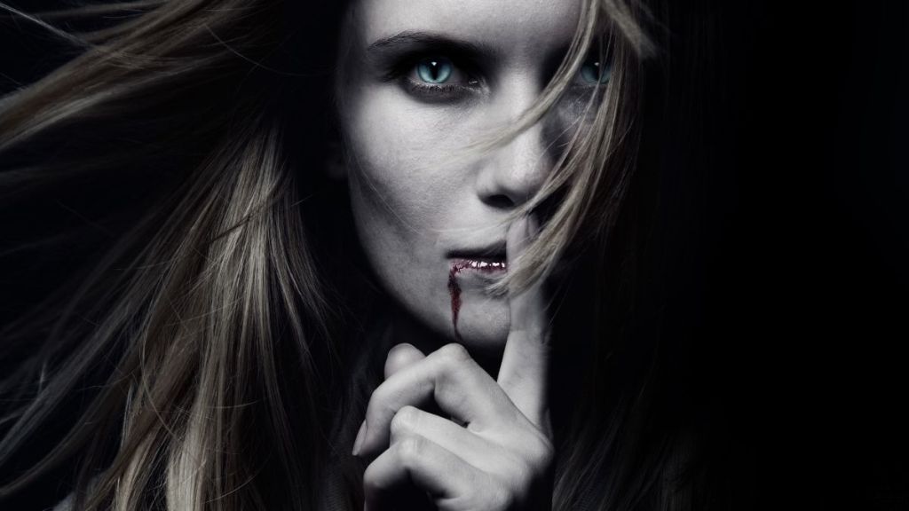 A blue-eyed woman who has blood dripping from her lips and a finger on her lips