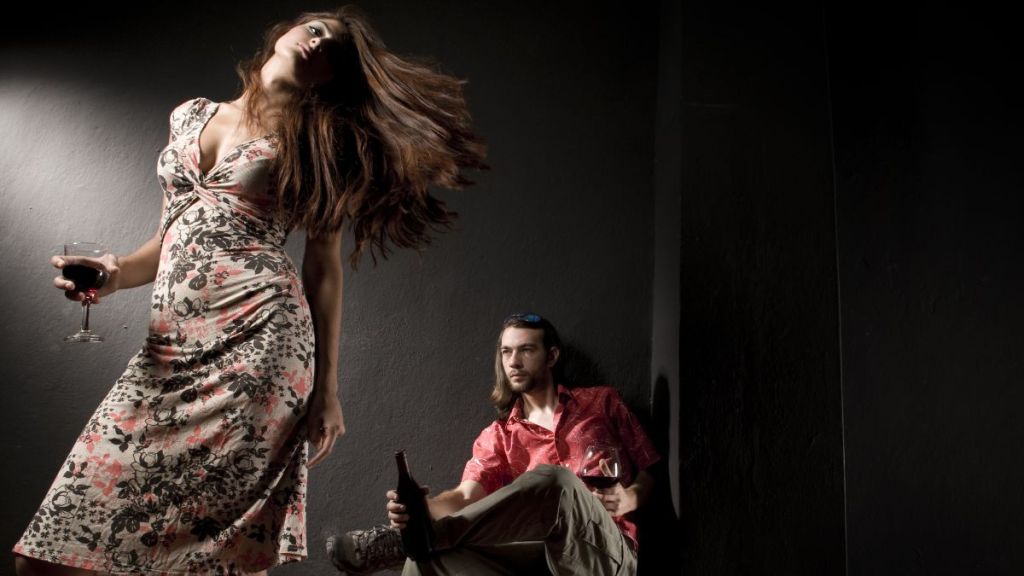 A woman standing with a wine glass in her hand and her head tilted to the side while a man sits diagonally behind her with a wine glass in one hand and wine bottle in the other